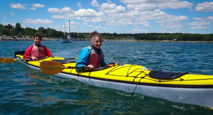 Two students sit in a yellow kayak floating on water and smile at the camera